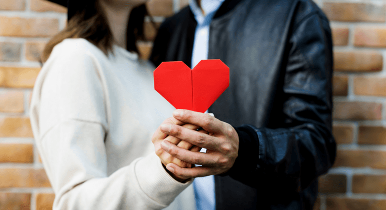 Man and woman stand while jointly holding up a red origami heart  